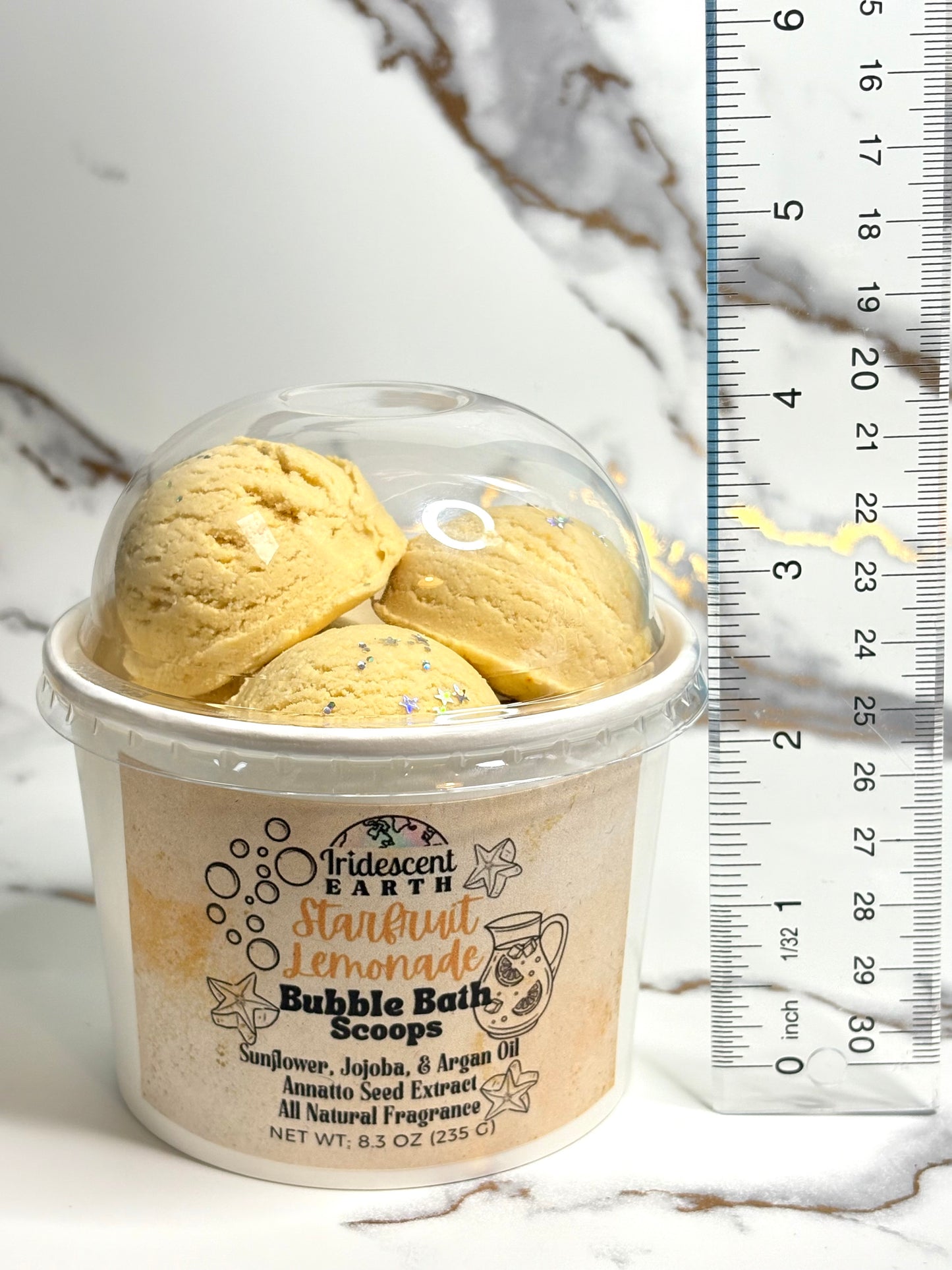 Container of Starfruit lemonade scented bubble bath scoops with a ruler next to it on a marble background
