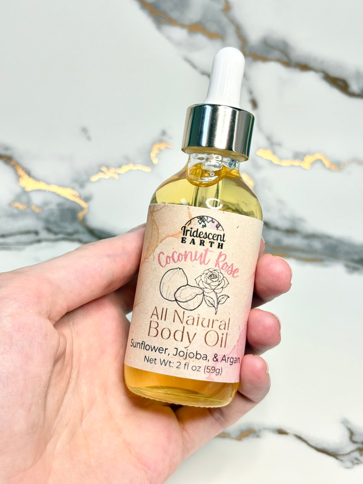 Hand holding a bottle of Iridescent Earth Coconut Rose All Natural Body Oil against a marble background.